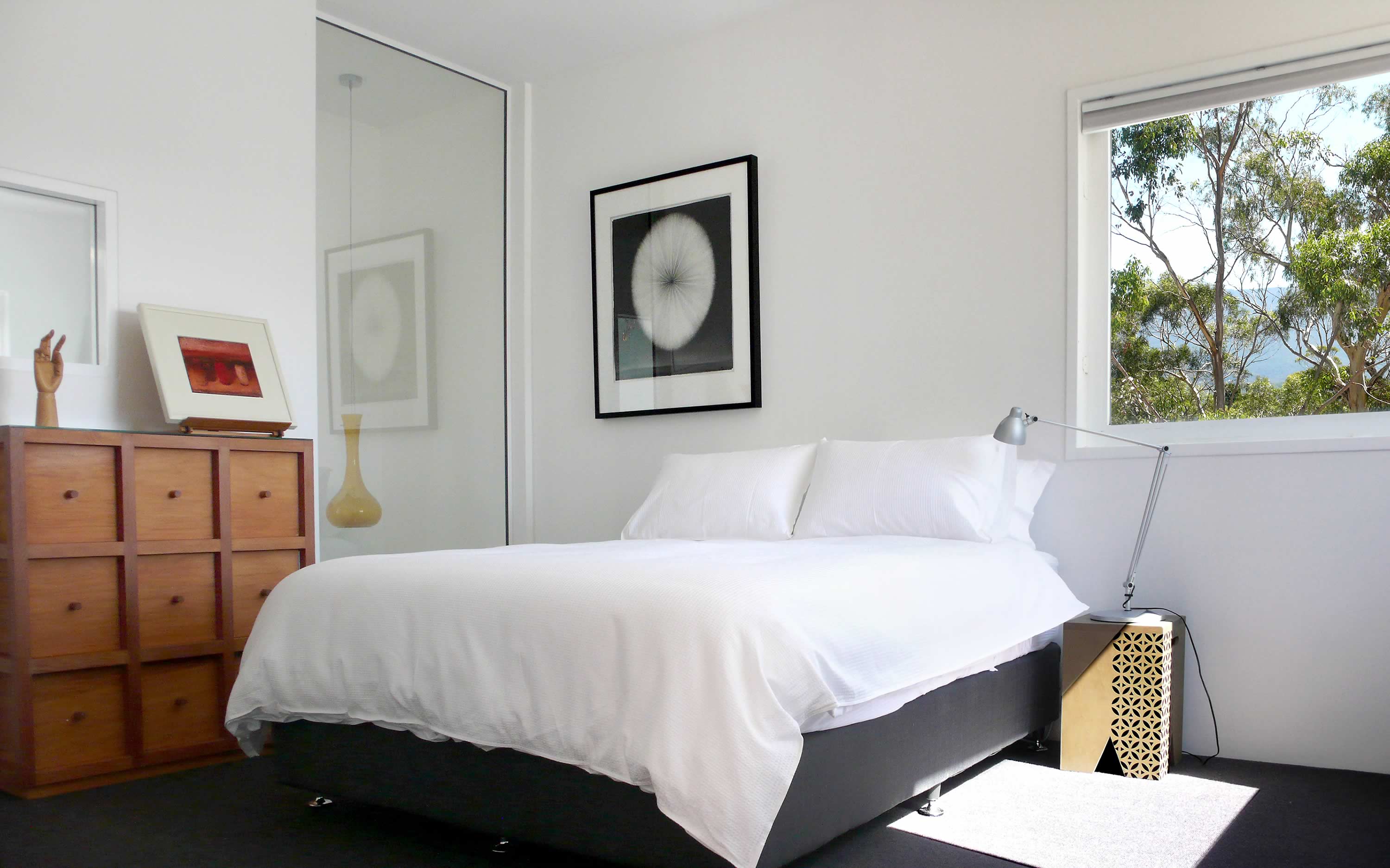 Photo of the OUTSIDE THE BOX bedroom showing a white-covered bed and art works, with eucalypt canopy visible through a window.