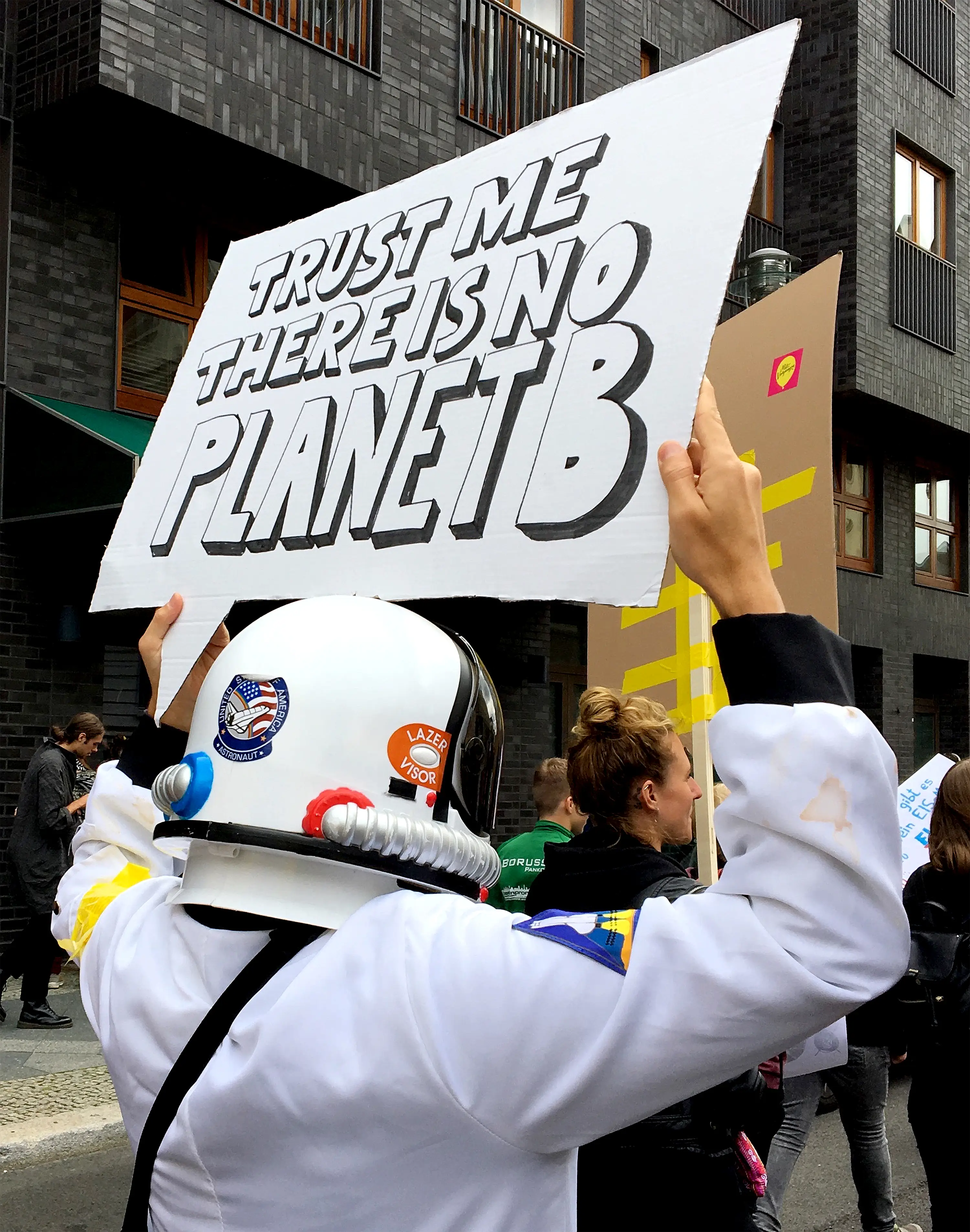 Picture of a person at a climate change rally wearing a spacesuit and holding a placard above their head reading ‘trust me there is no Planet B’.