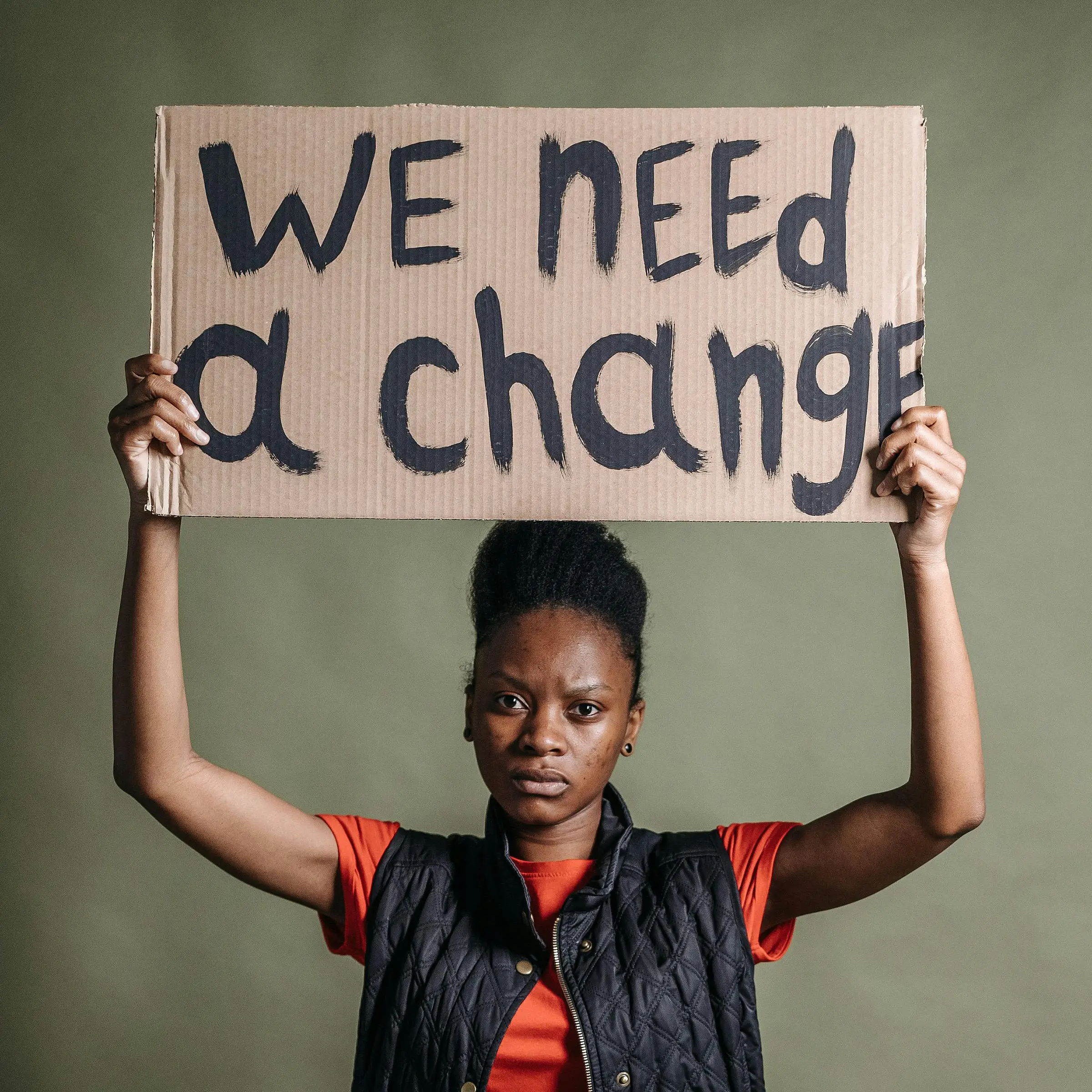 Picture of a woman wearing an orange t-shirt and black vest holding a cardboard sign above her head that reads ‘we need a change’.