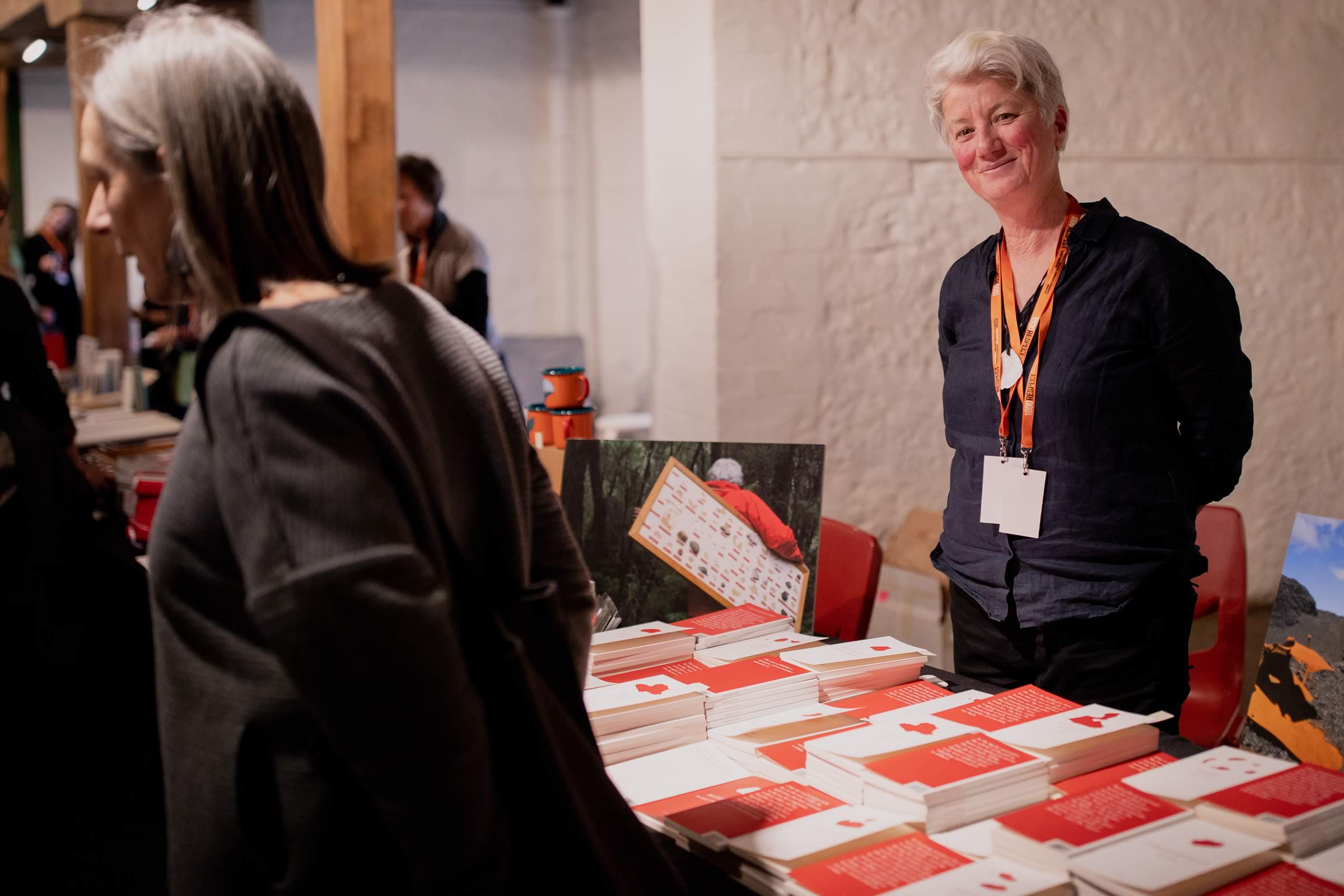 Photo of a smiling woman stallholder (event organiser, Margaret Woodward) standing behind a display of red and white covered books, with a woman walking past the stall in the foreground. Photo: Rosie Hastie for The People’s Library 2018.