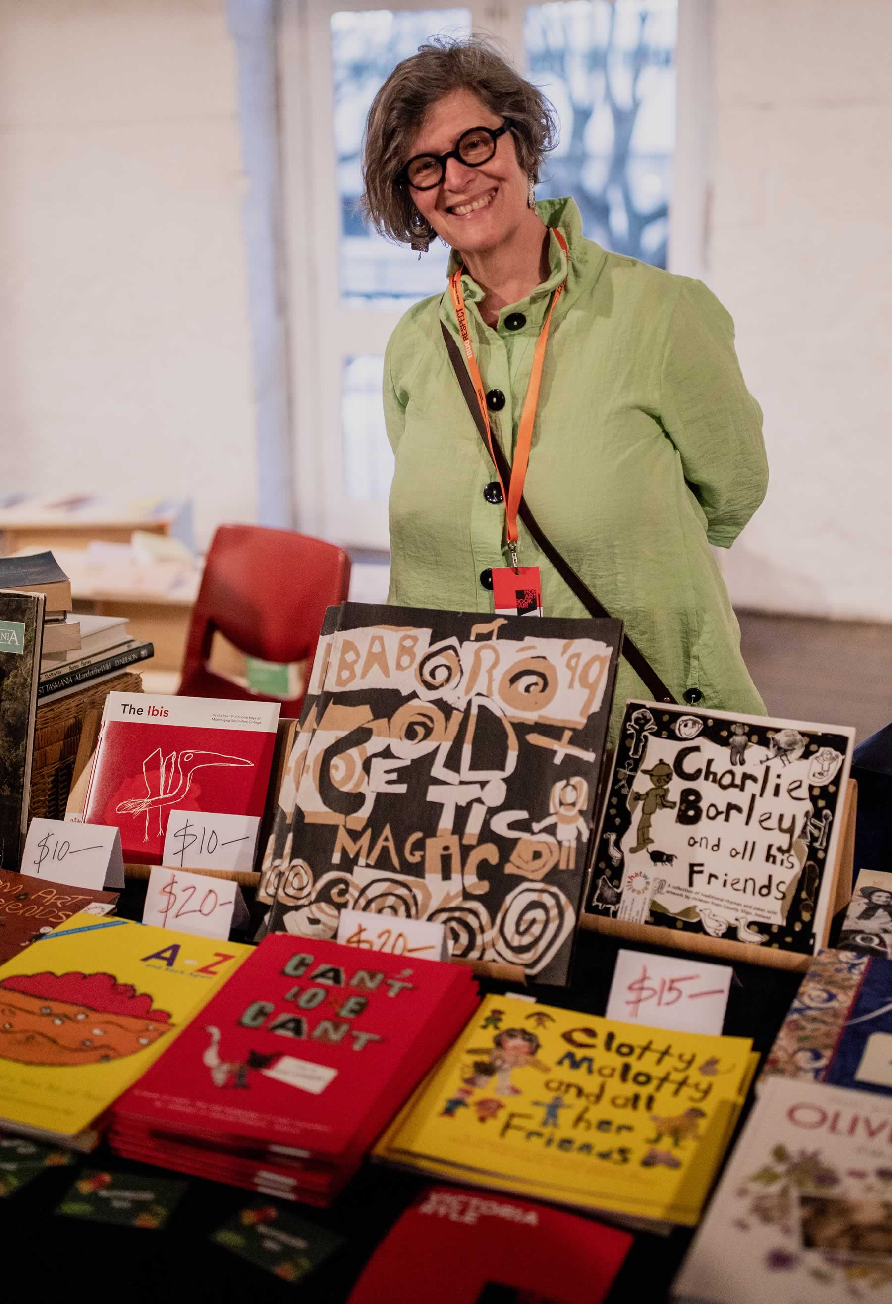 Photo of a smiling woman stallholder behind a display of a number of colourful children’s books. Photo: Rosie Hastie for The People’s Library 2018.