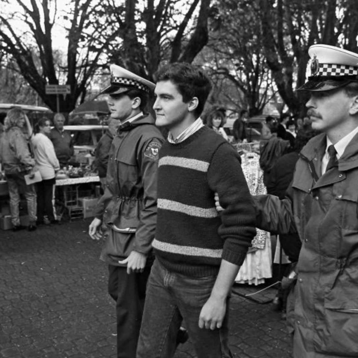 Black and white photo of gay activist Rodney Croome being escorted from the 1998 Salamanca Market gay rights protests by a police officer.
