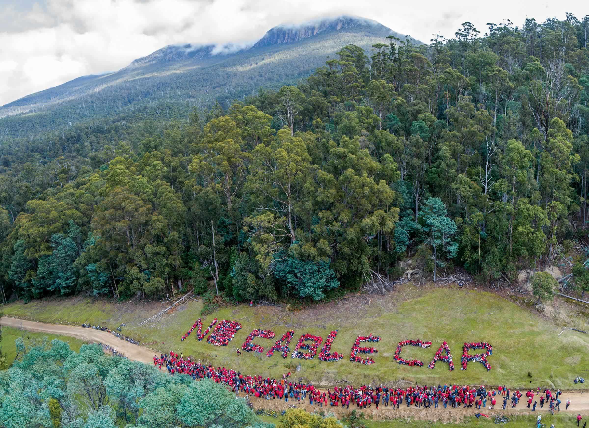 Photo from the air of a protest in which people dressed in red form the words “No Cable Car” against a grass hillside, with the forest and mountain in the background.