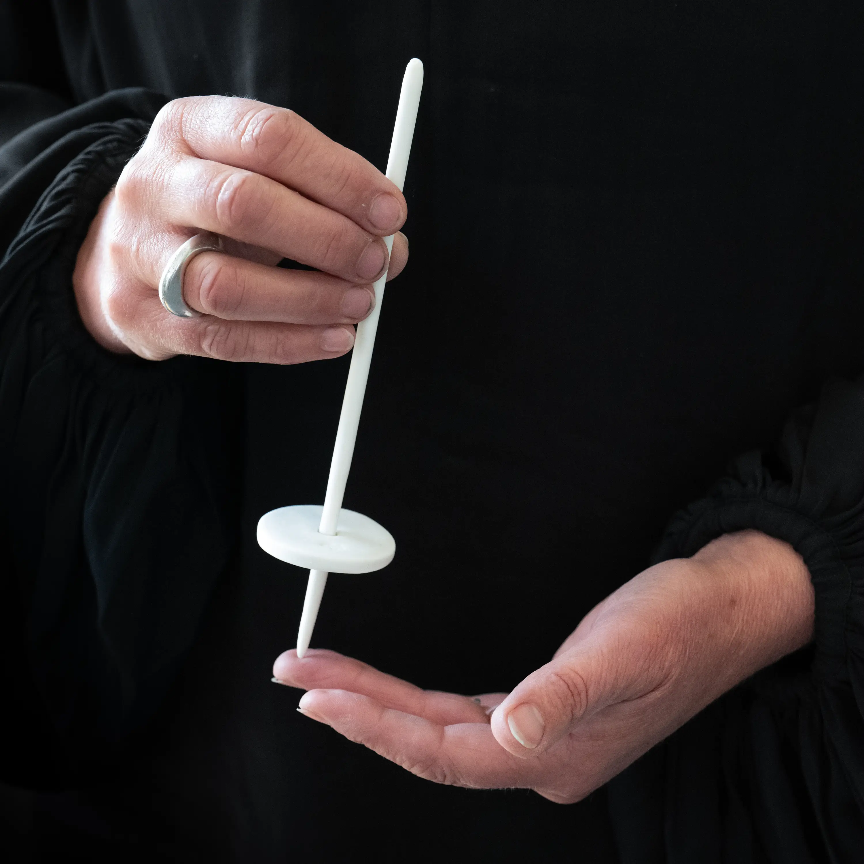 Image of a woman’s hands against a black background holding a delicate needle shaped white ceramic object.