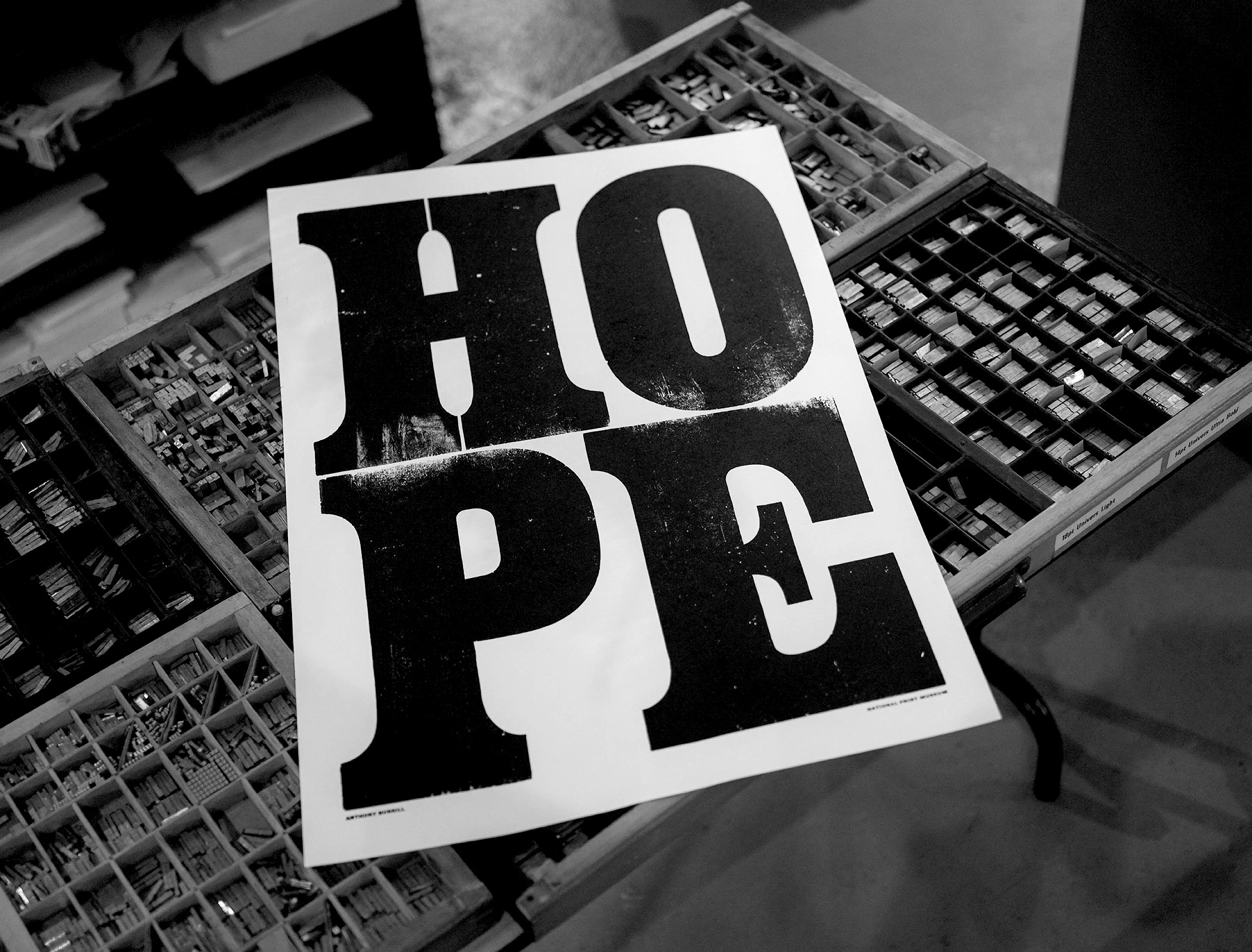 An image of a black and white poster with large capital letter type spelling out the word HOPE. The poster is sitting on top of a tray with individual compartments filled with small metal pieces of type.