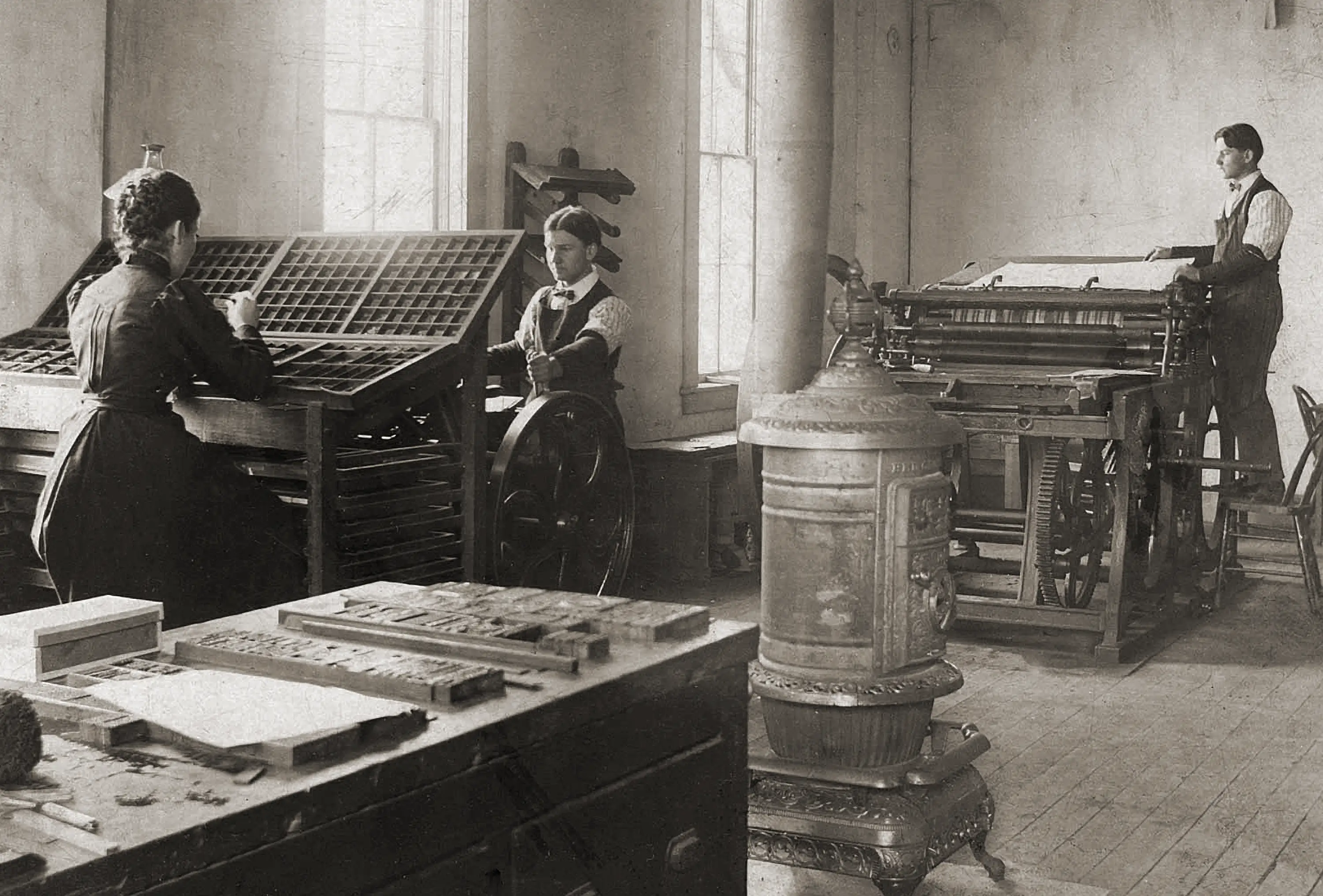 A sepia toned image titled The First Print Shop c.1885&ndash;90. A women is working at a compositors desk sorting small metal type and two men are operating large letterpress machines.