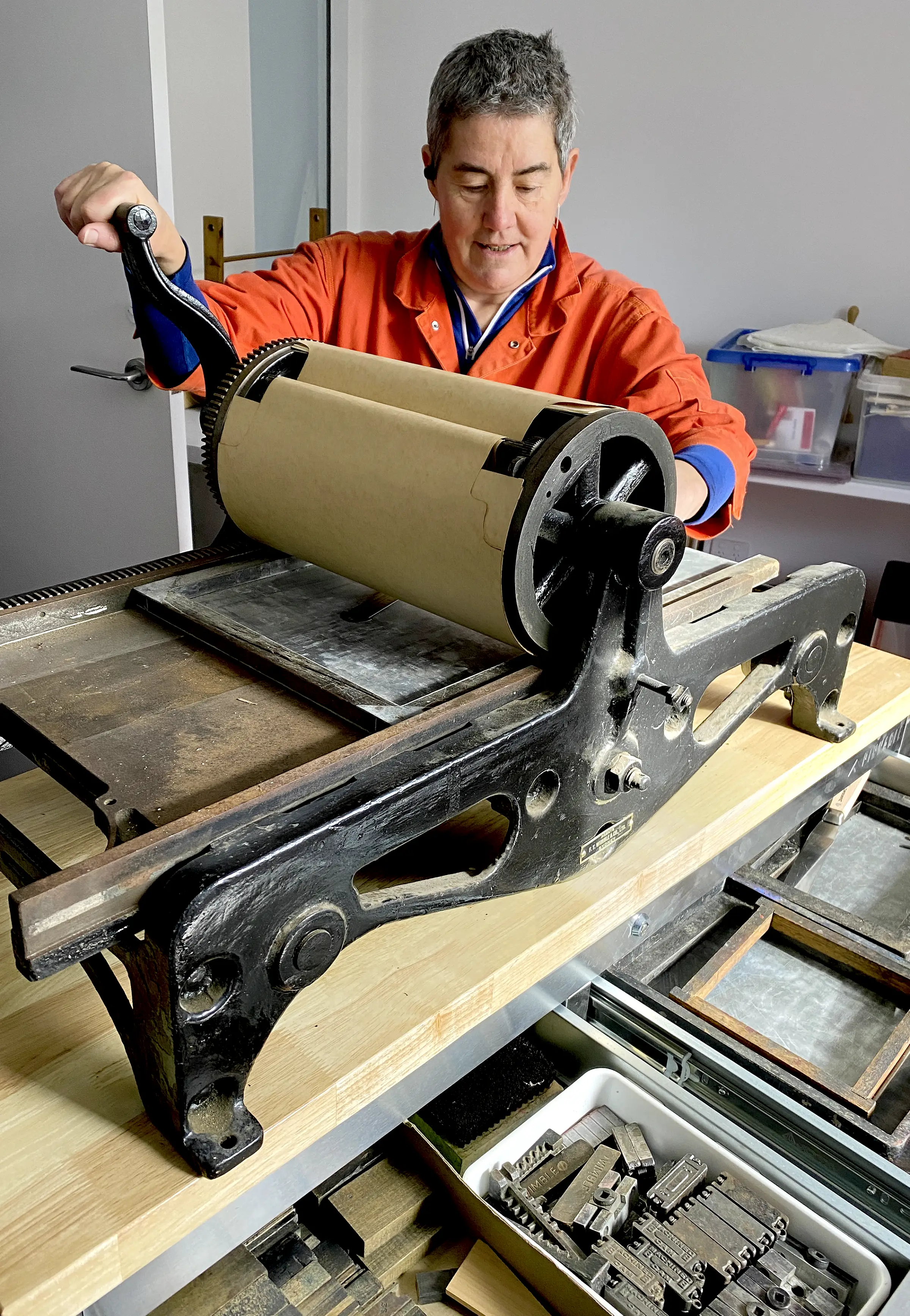An image of a woman dressed in a bright orange set of overalls operating the roller on a small letterpress.
