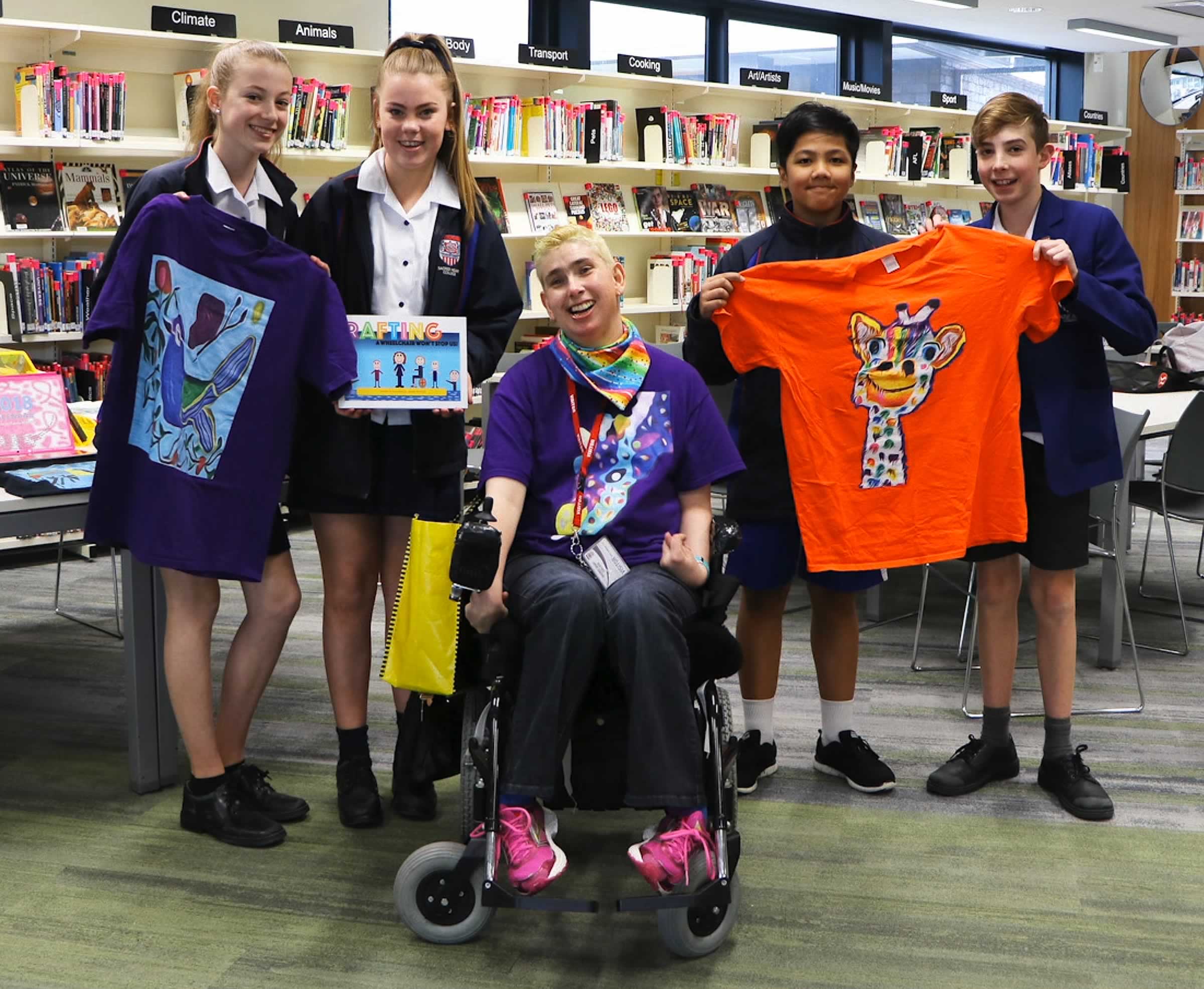 Photo of Janelle McMillan with four school children holding some of her t-shirts and books, with bookshelves behind them — all smiling.