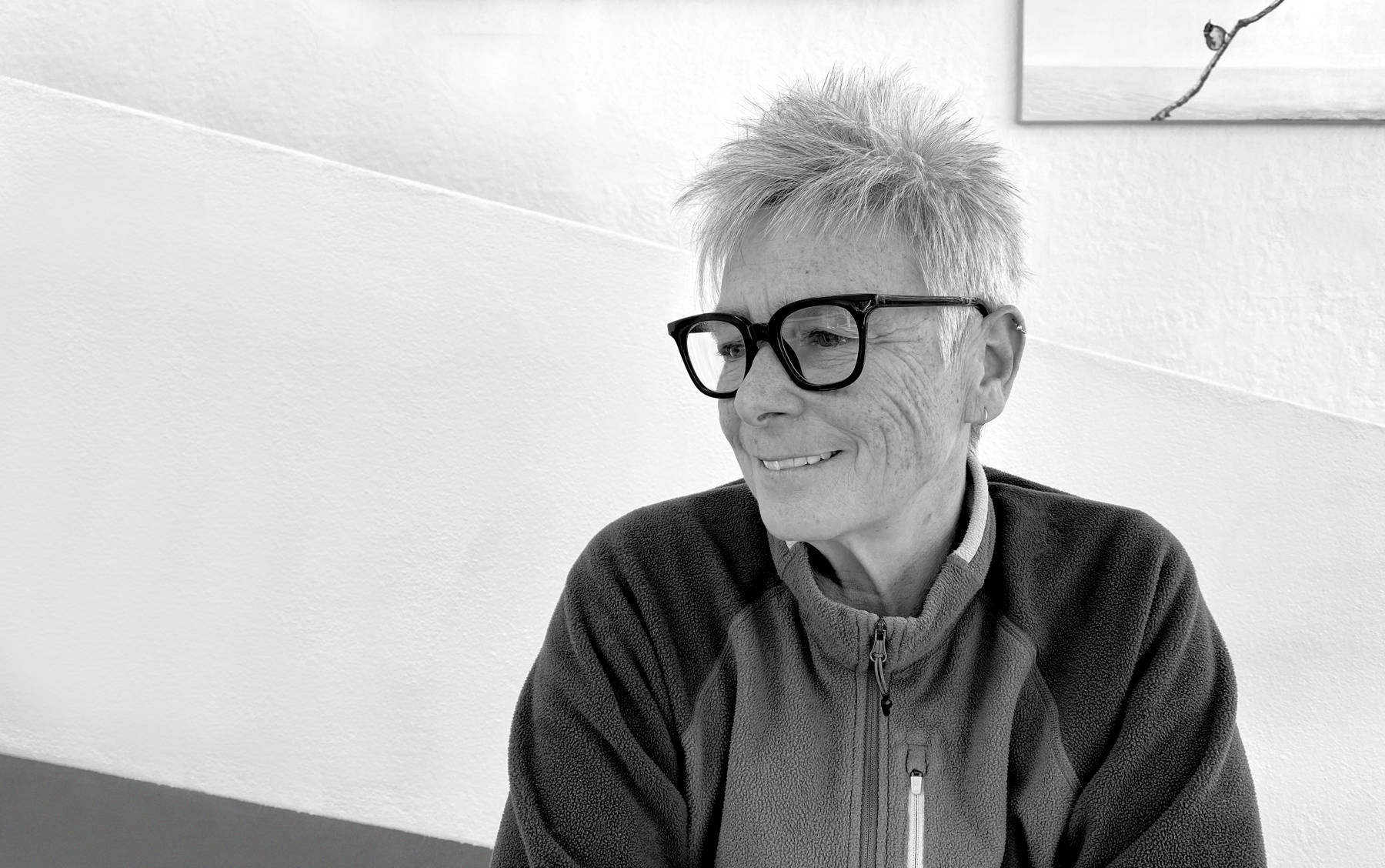 A black and white portrait image of Tracey Diggins with her face turned slightly in profile. She has short, spiky grey hair and is wearing large-framed black reading glasses.