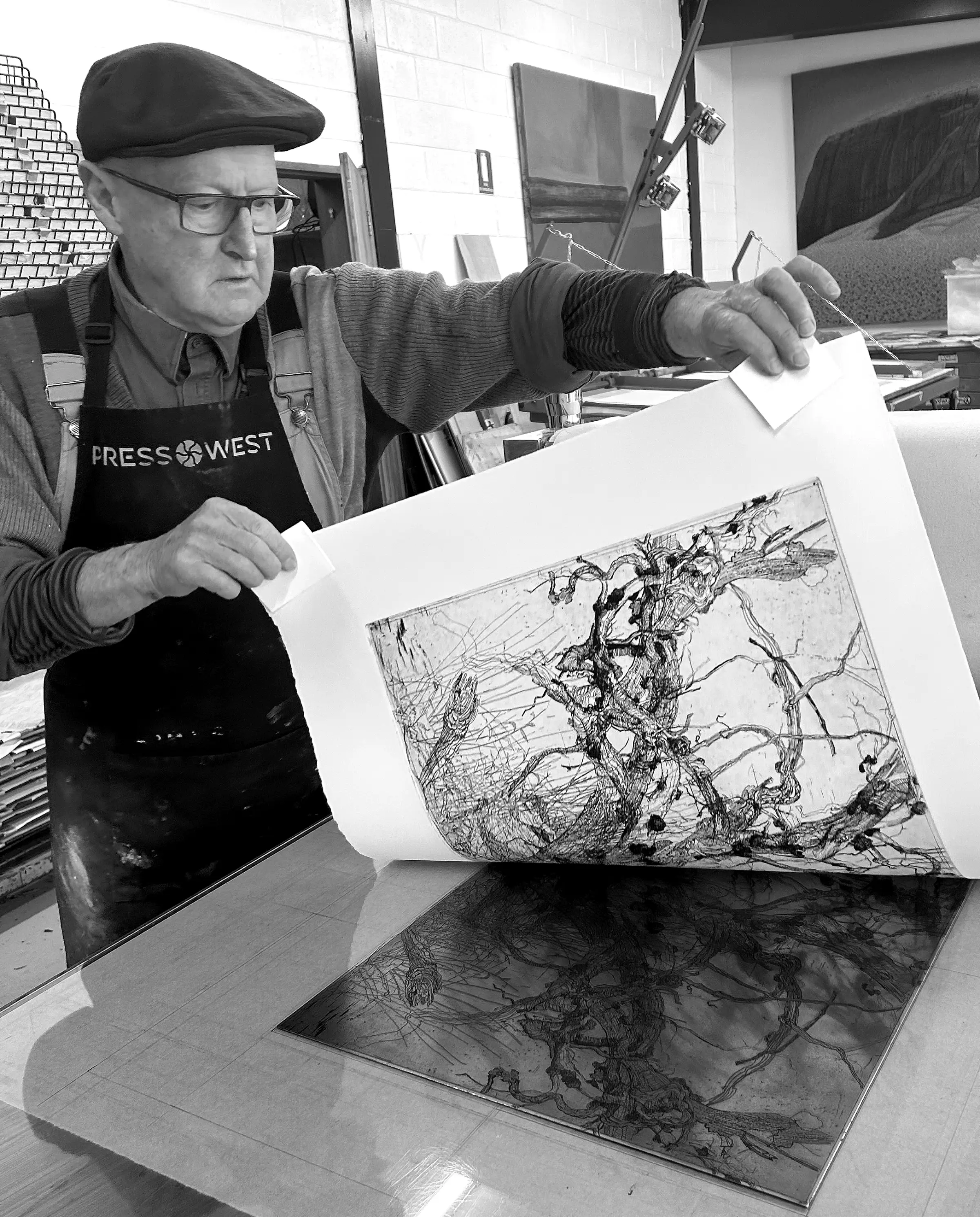 A black and white portrait of a man wearing a peaked cap, glasses and artist&rsquo;s apron. He is lifting an etching off a copper plate.