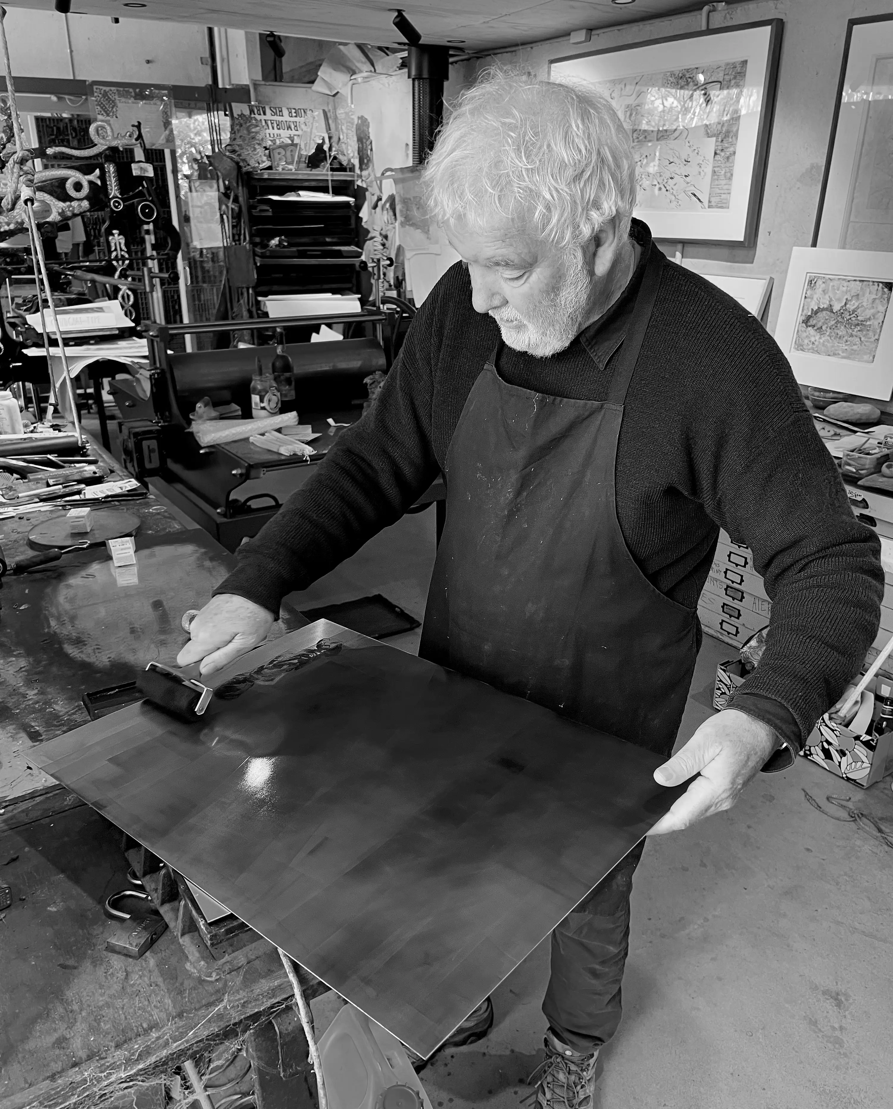A black and white portrait of a man with grey hair in an artist studio. He is using a small roller to ink up a copper etching plate.