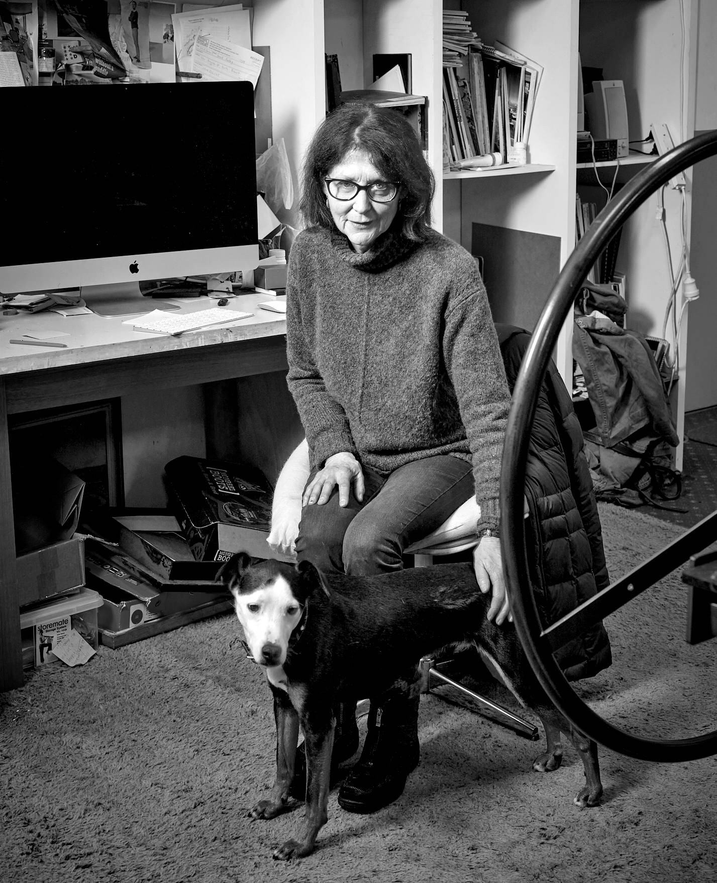 A black and white portrait of artist Helen Wright sitting in a chair in her studio. She is patting her small white-faced rescue dog Mr Dexter. An imposingly large wheel of a lithograph press is visible in the foreground.