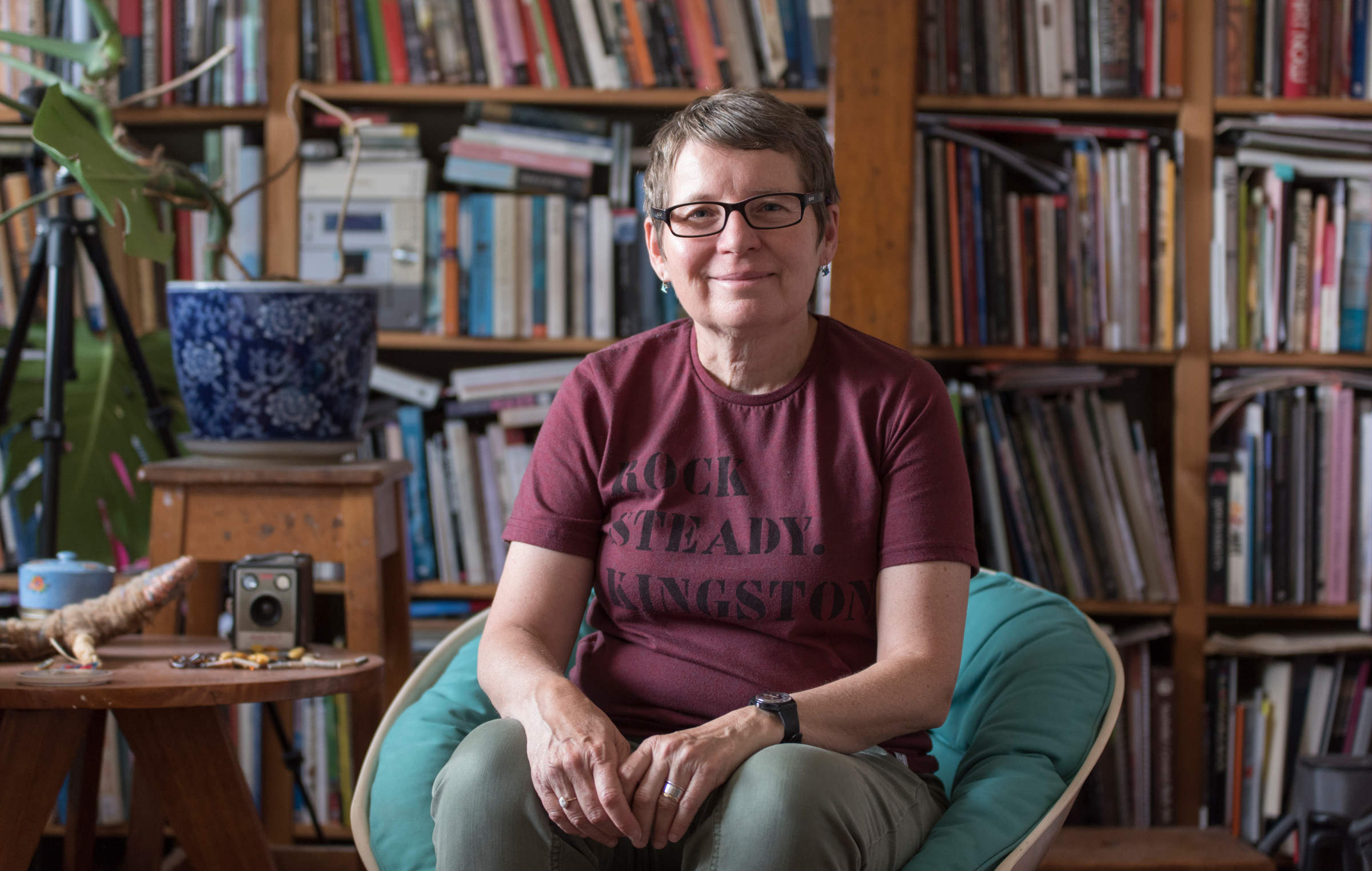 Portrait of artist Julie Gough by photographer Lucy Parakhina. Julie is seated amongst a backdrop of her extensive library of books and smiling at the camera.