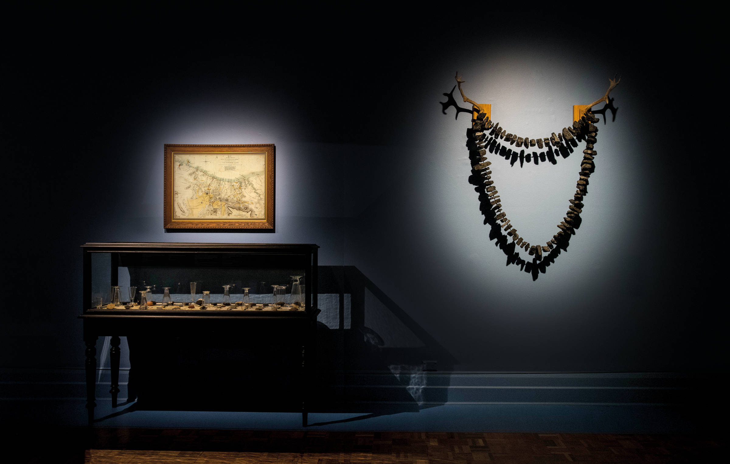 TENSE PAST exhibition image by Alistair Bett depicting two artworks. On the left-hand side of the image is a multi-form installation work titled, ‘North Down’ (2019). This work incorporates a colonial era cabinet filled with various Tasmanian Aboriginal stone tools collected from the Northdown, colonial property in Tasmania and a collection of Victorian era apothecary measuring glasses. Hanging above the cabinet is an historic map drafted in 1828 by Henry Hellyer (1790–1832) titled, ‘Map of the Interior Discoveries made by the Van Diemen’s Land Company (in exploring their location) Van Diemen’s Land 1828’. Inside the cabinet (but not visible), is a sketchbook of watercolour drawings by Thomas Scott titled, Sketches, ‘Van Diemen’s Land 1822–1847’, with the inscription ‘From North Down Hill to the Beach at Port Sorell…’ On the right-hand side of the image, hanging on the wall is a sculptural work titled, ‘Killymoon’ (2008). This large 3-D work is representative of an over-sized necklace made from lumps of Fingal Valley coal, hanging from fallow deer antlers on wooden fixing points.