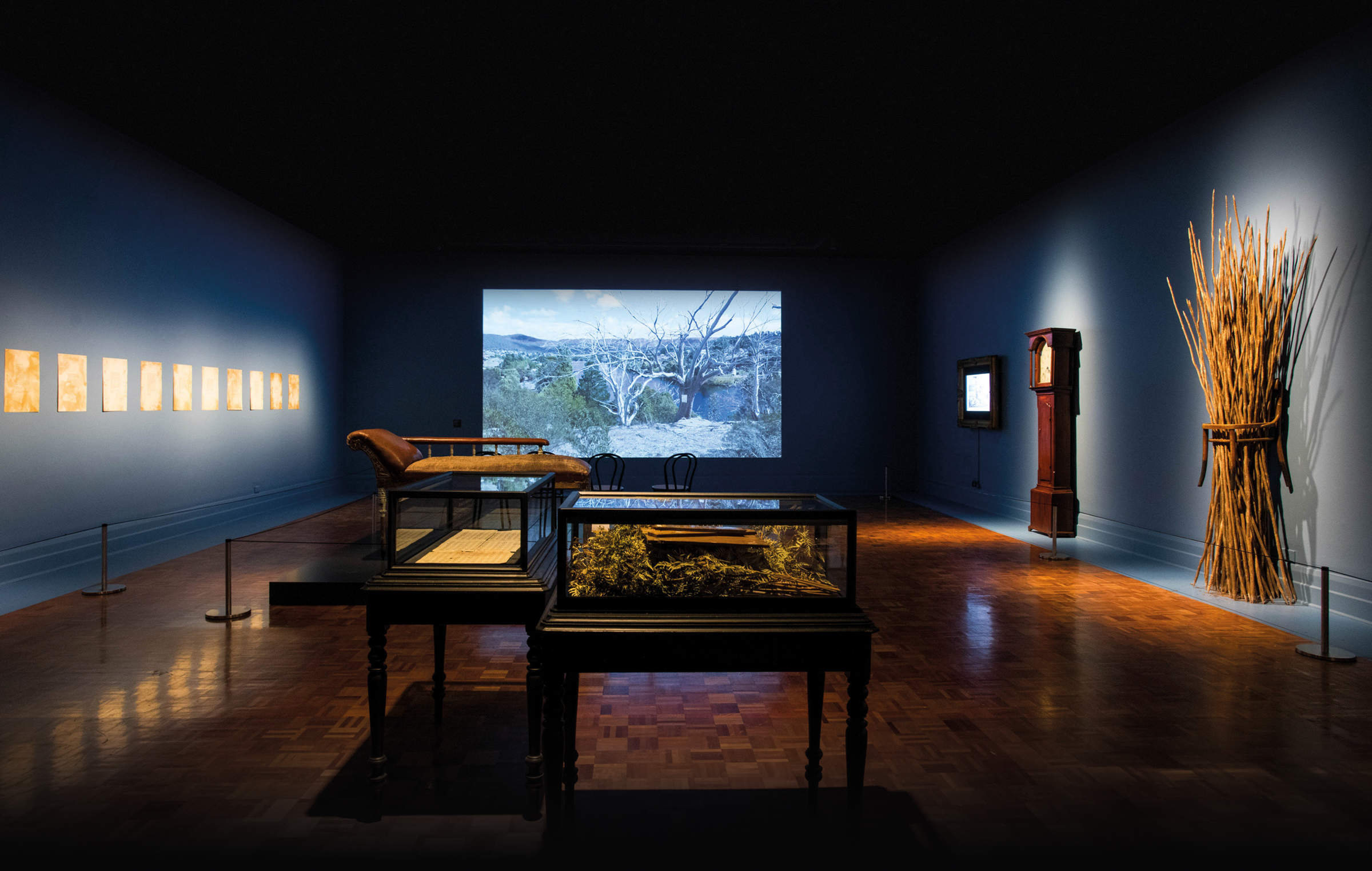 TENSE PAST exhibition image by Alistair Bett depicting a room containing multiple artworks and historical artefacts. On the left-hand wall is a work titled, ‘Hunting Ground (Haunted) Van Diemen’s Land’ (2016) consisting of 10 etching and acrylic silkscreen prints. On the front wall is a video work showing some dead trees in the landscape titled, ‘Hunting Ground (Haunted) Van Diemen’s Land’ (2016–17). A smaller video work sits on the right-hand wall titled, ‘Hunting Ground (Pastoral) Van Diemen’s Land’ (2016–17). Next to the screen on the same wall is a mahogany and painted copper Longcase clock c.1820. Alongside the clock is a sculptural work titled, ‘Some Tasmanian Aboriginal Children Living with Non-Aboriginal People before 1840’ (2008) consisting of a found chair with burnt tea tree sticks with names carved on them. In the centre of the room is a Sofa table c.1850 made from Huon pine, Australian red cedar and non-native white pine. The final work titled, ‘Luna riabi (song)’ (2019) is an audio work with music score referencing ‘Song of the Aborigines’ 1856 by Mrs Maria Logan (1808–1886).