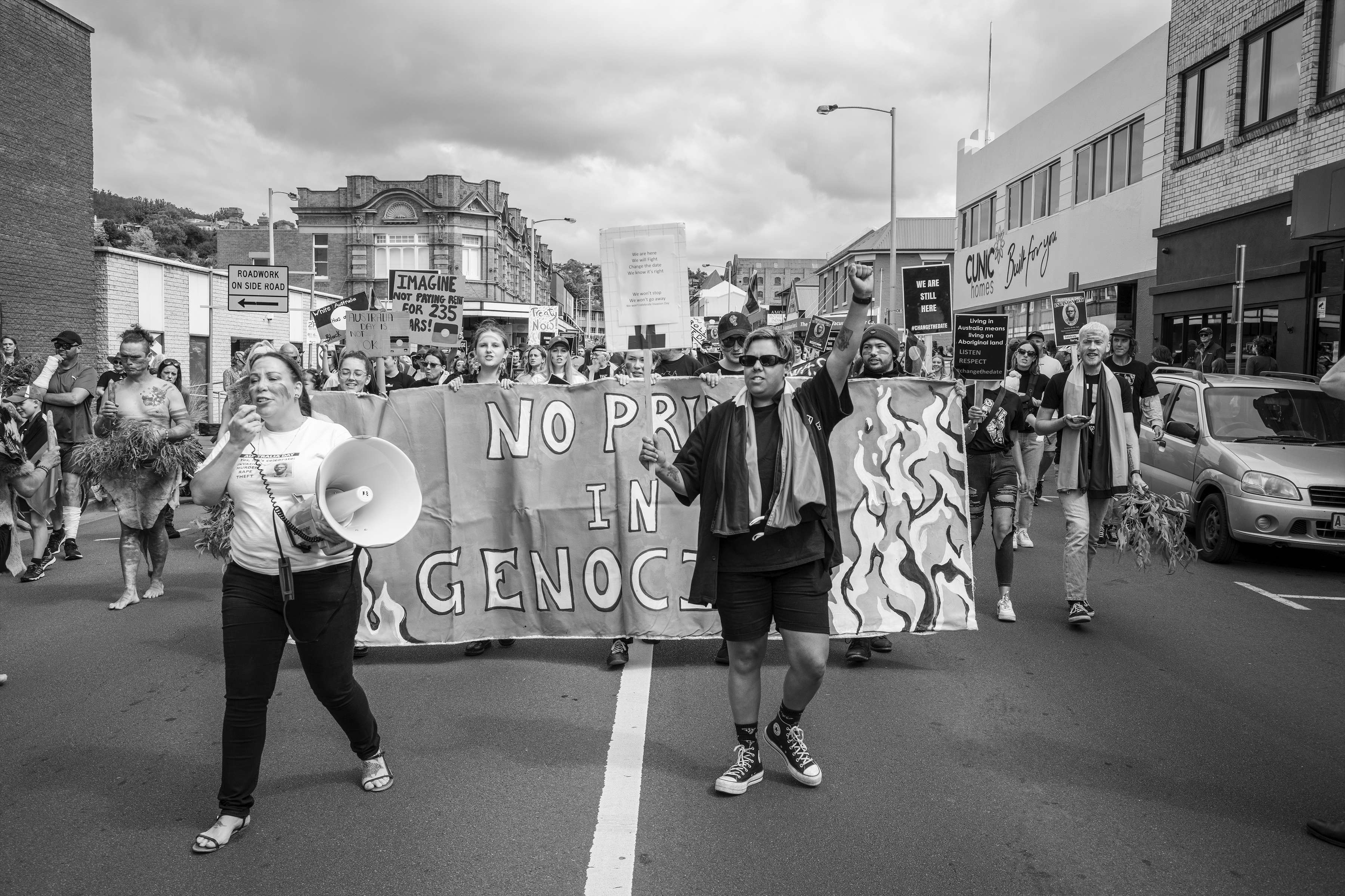 An image of a group of Aboriginal protesters walking and chanting down Elizabeth Street in the city of nipaluna/Hobart. The lead protestor has a megaphone. The main protest sign reads No pride in genocide.