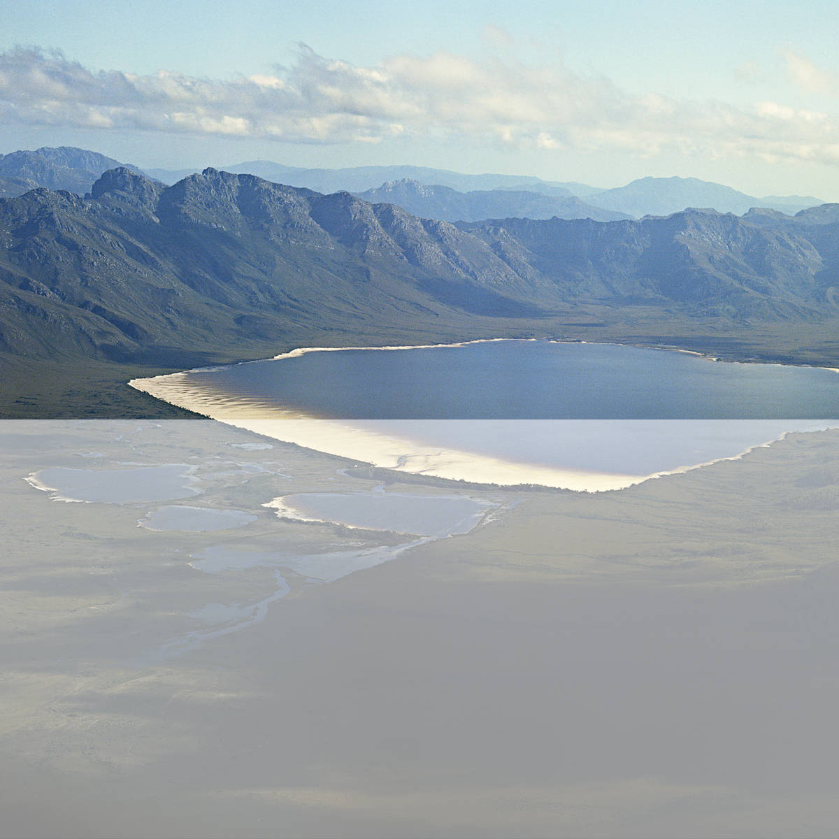 Photograph of the original Lake Pedder by Wilf Elvy taken from a light plane in 1972–73 prior to inundation by the Huon-Serpentine impoundment waters. The rugged Frankland Range is in the background and the three kilometre long, stunning pink quartzite beach sparkles in the foreground.