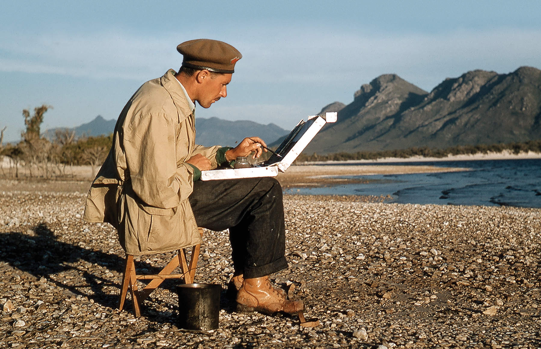 Photograph of artist Max Angus by fellow artist Patricia Giles captures Max as a young man wearing his trademark beret and seated on the shore of Lake Pedder, painting en plein air.