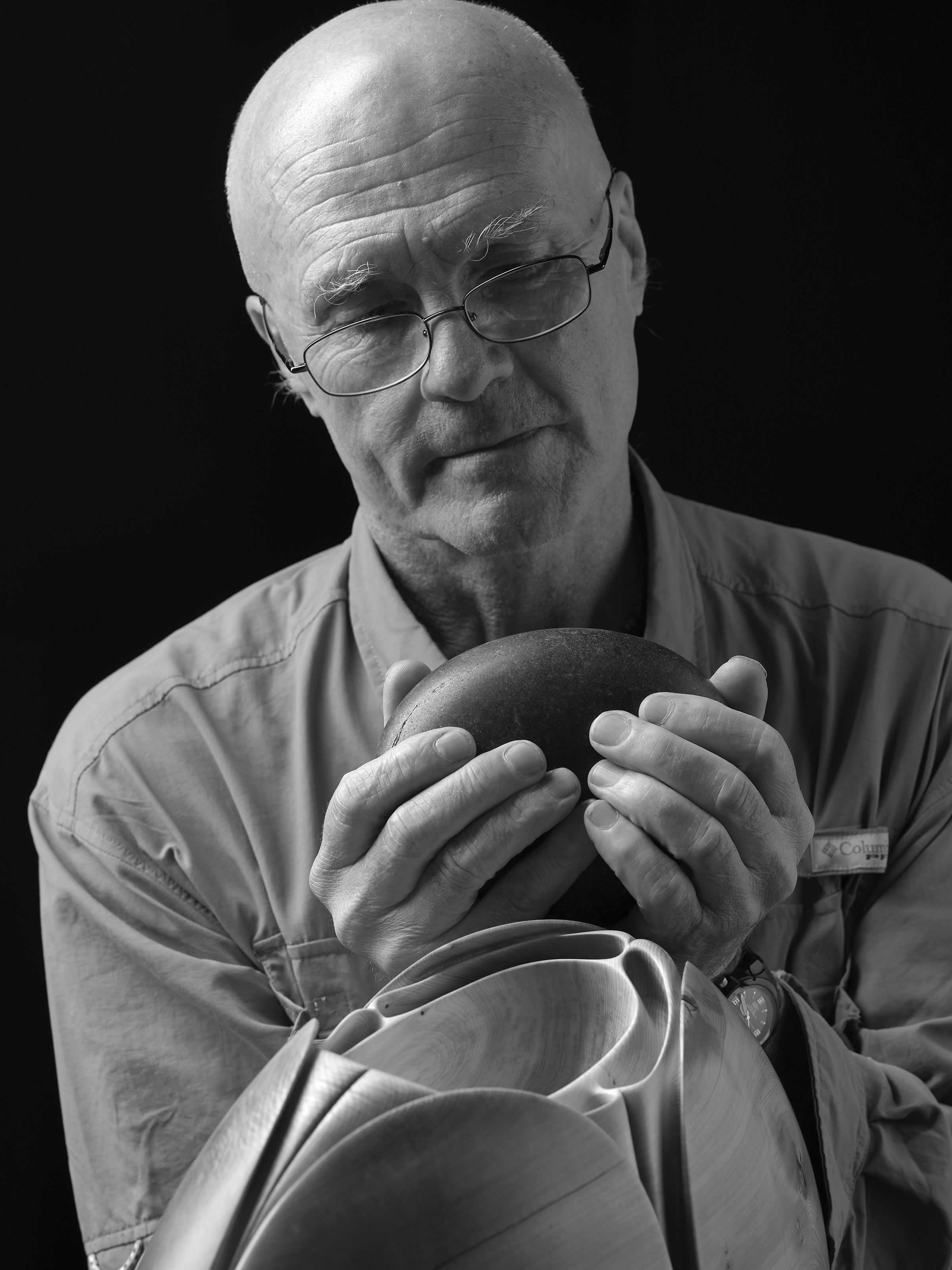 Black and white portrait photo of Peter Adams holding a black rock with a carved wooden sculpture in the foreground. Photo: Peter Whyte.