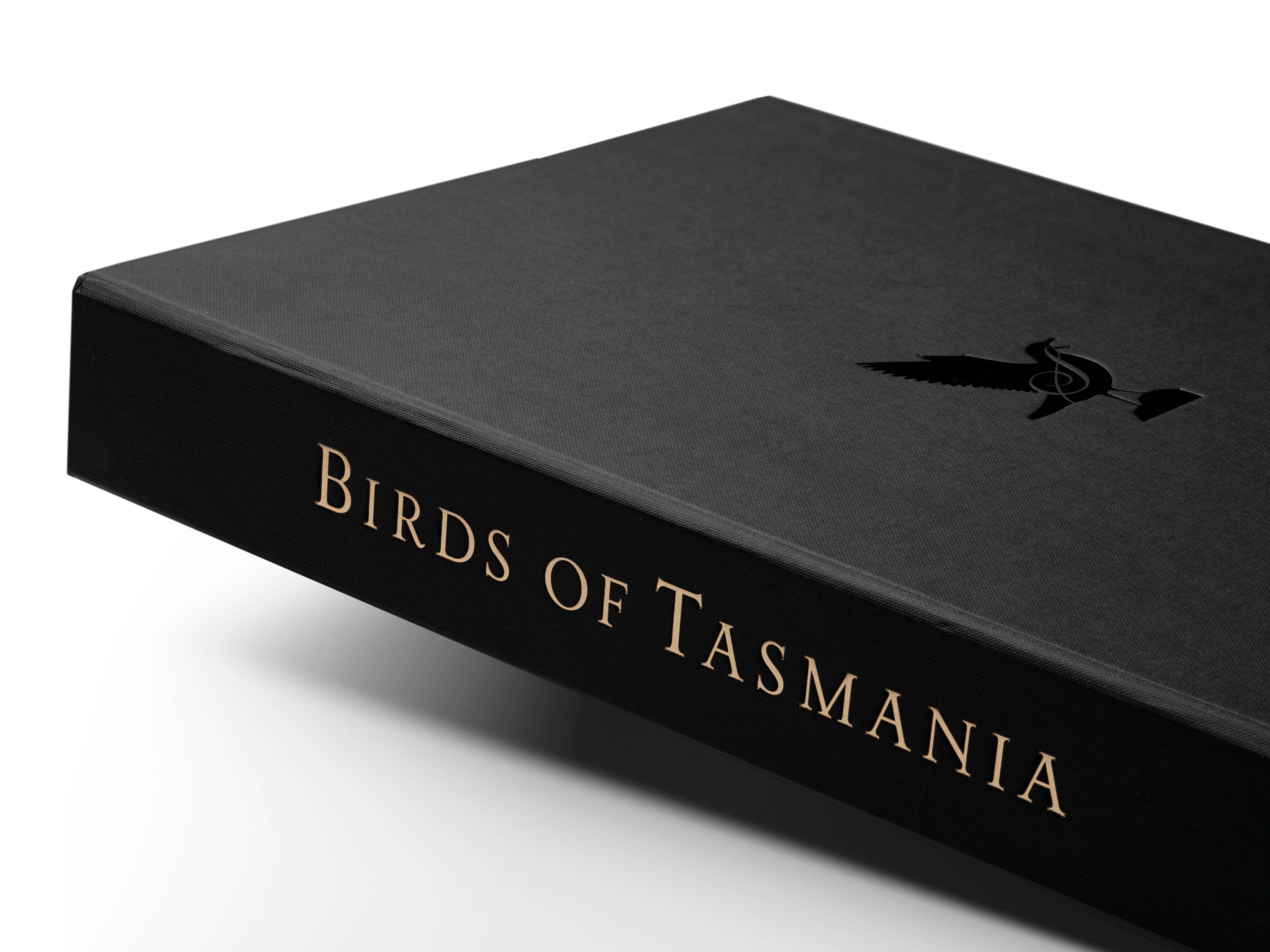 A close-up of a section of the ‘Birds of Tasmania’ by Susan Lester clamshell presentation box, showing gold foil titling and a black embossed symbol of a bird with the letter ‘S’ in a script font.