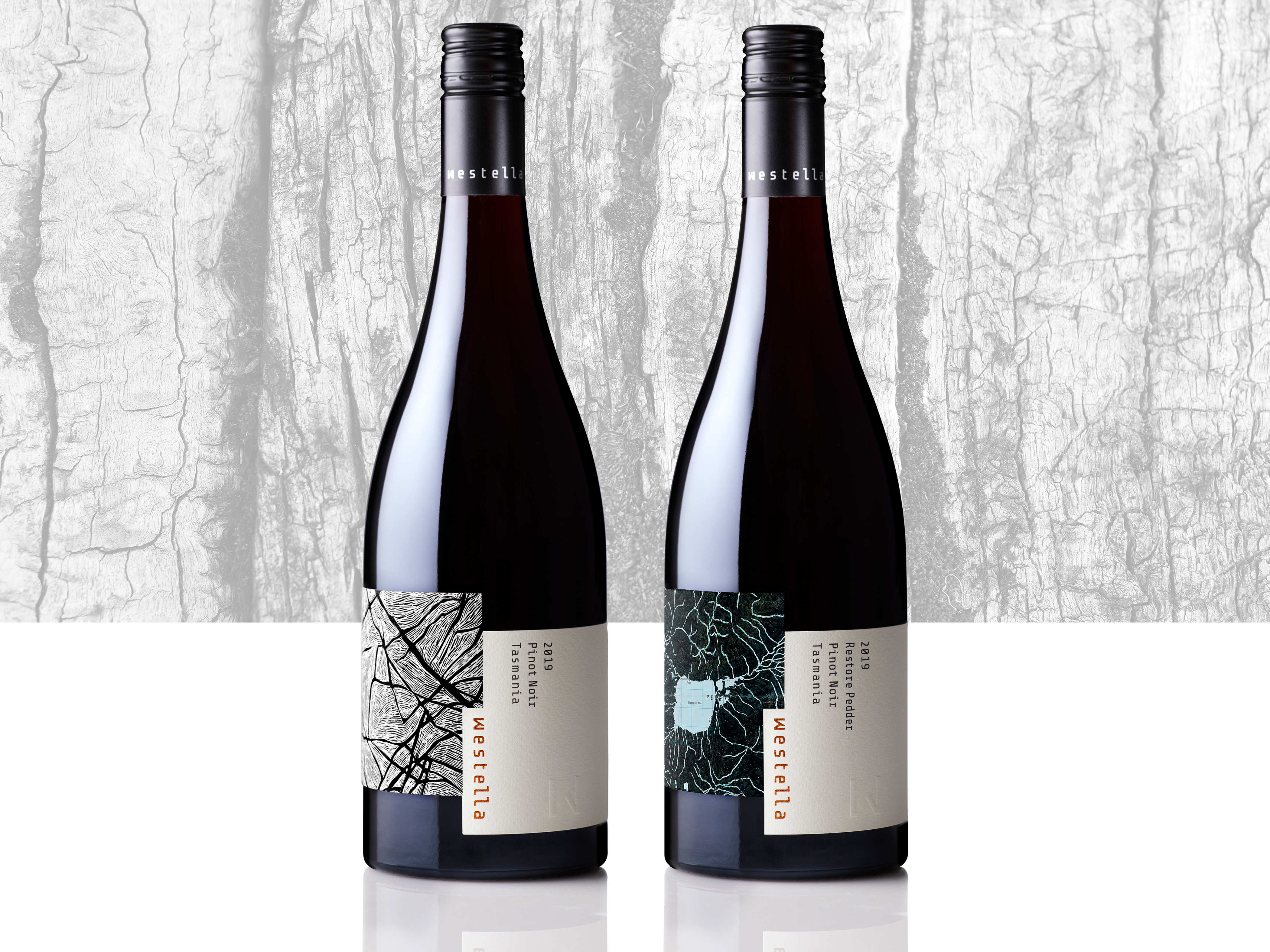 Two bottles of 2019 vintage Westella Pinot Noir sitting side by side against a backdrop of tree bark. Both bottles feature labels with artwork by Sue Lovegrove. The bottle on the left has the work titled, ‘The long walk, Skullbone Plains, Tasmania’ (2014) which is a black and white abstract pattern. The bottle on the right is a special release label featuring a detail from ‘Mapping the invisible, [reconstructed map]’ (2021) that Sue created for the water[shed] exhibition.