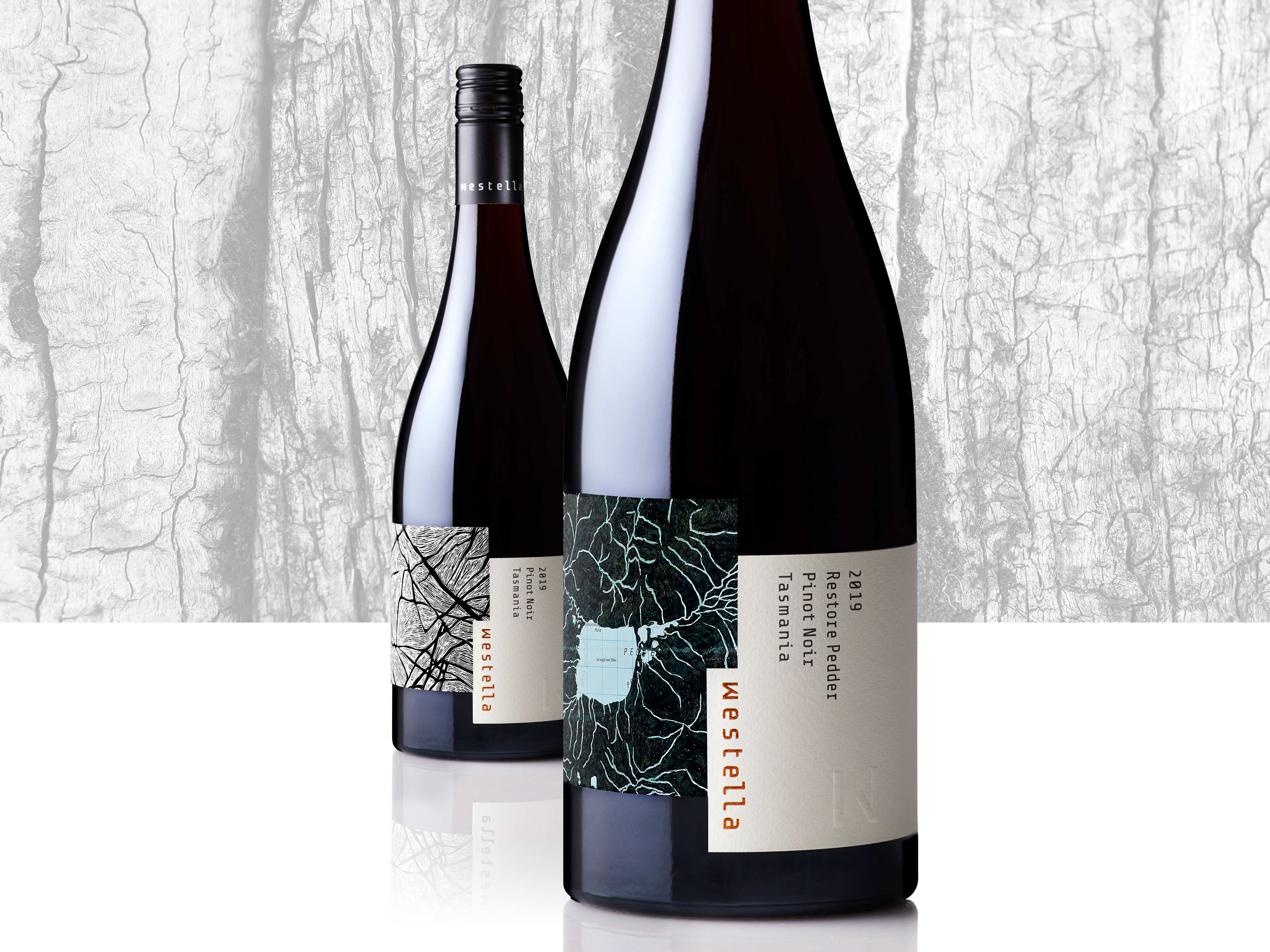 Two bottles of 2019 vintage Westella Pinot Noir sitting one behind the other against a backdrop of tree bark. Both bottles feature labels with artwork by Sue Lovegrove. The bottle towards the back has the work titled, ‘The long walk, Skullbone Plains, Tasmania’ (2014) which is a black and white abstract pattern. The bottle at the front is a special release label featuring a detail from ‘Mapping the invisible, [reconstructed map]’ (2021) that Sue created for the water[shed] exhibition.