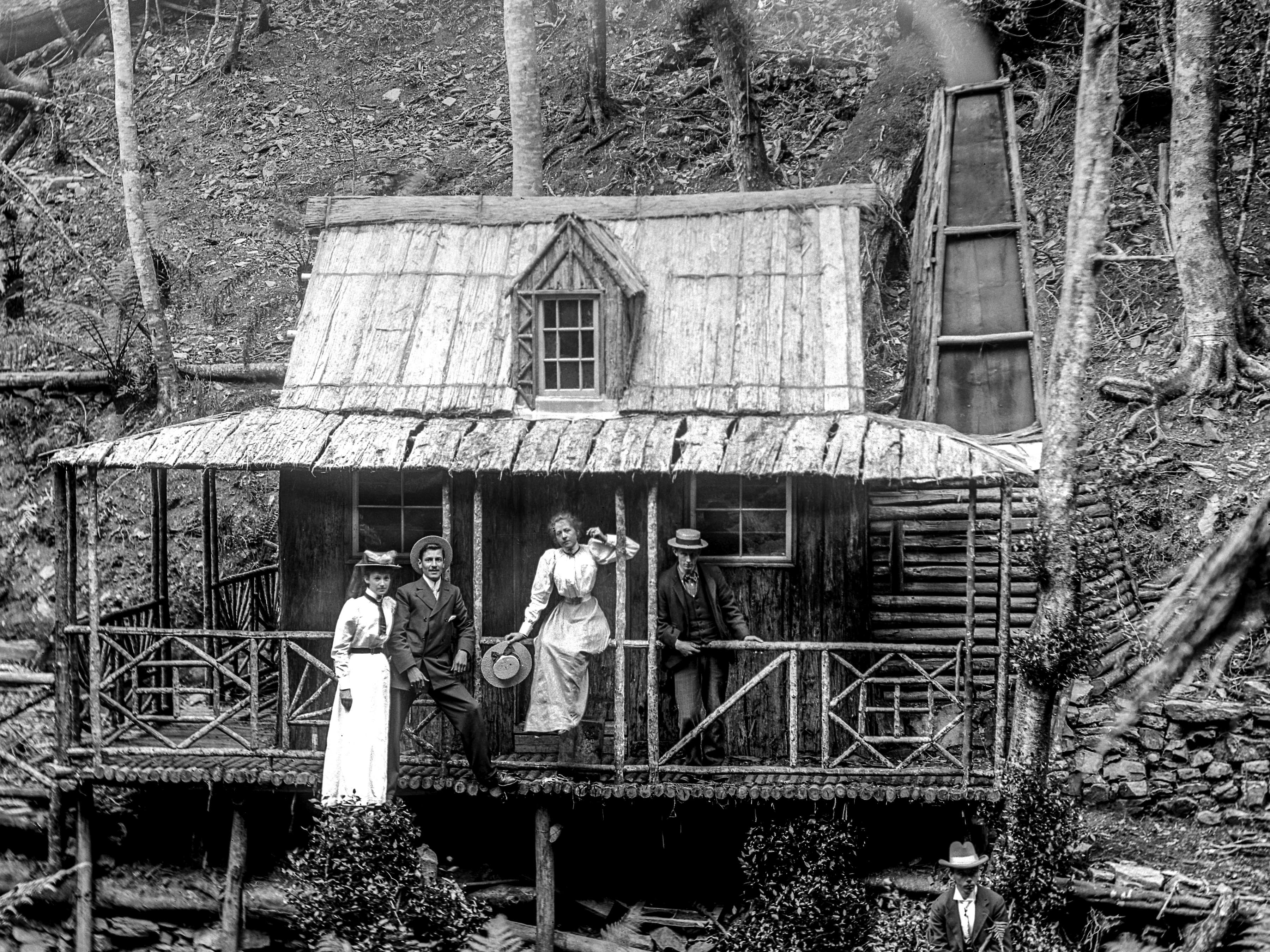 A black and white photograph of Wellington Hut nestled amongst the forest on the slopes of the mountain. Two couples are on the verandah dressed in their Sunday best and smoke from the hut's fireplace is gently billowing from the chimney.