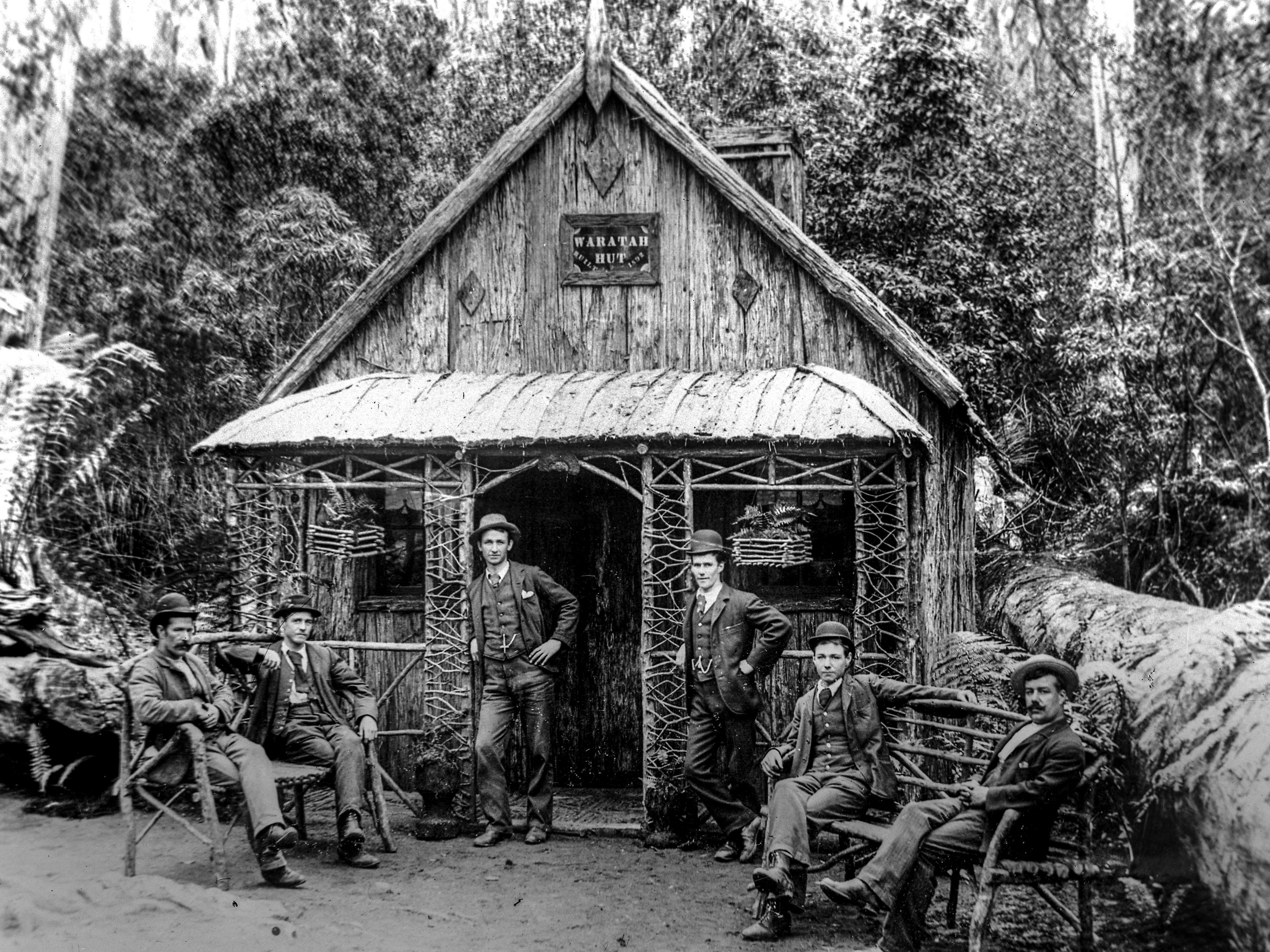 A black and white photograph of Waratah Hut nestled amongst the forest on the slopes of the mountain. Six men, some wearing bowler hats, are posing for the photographer either sitting on bench seats or standing leaning on the ornate verandah posts.