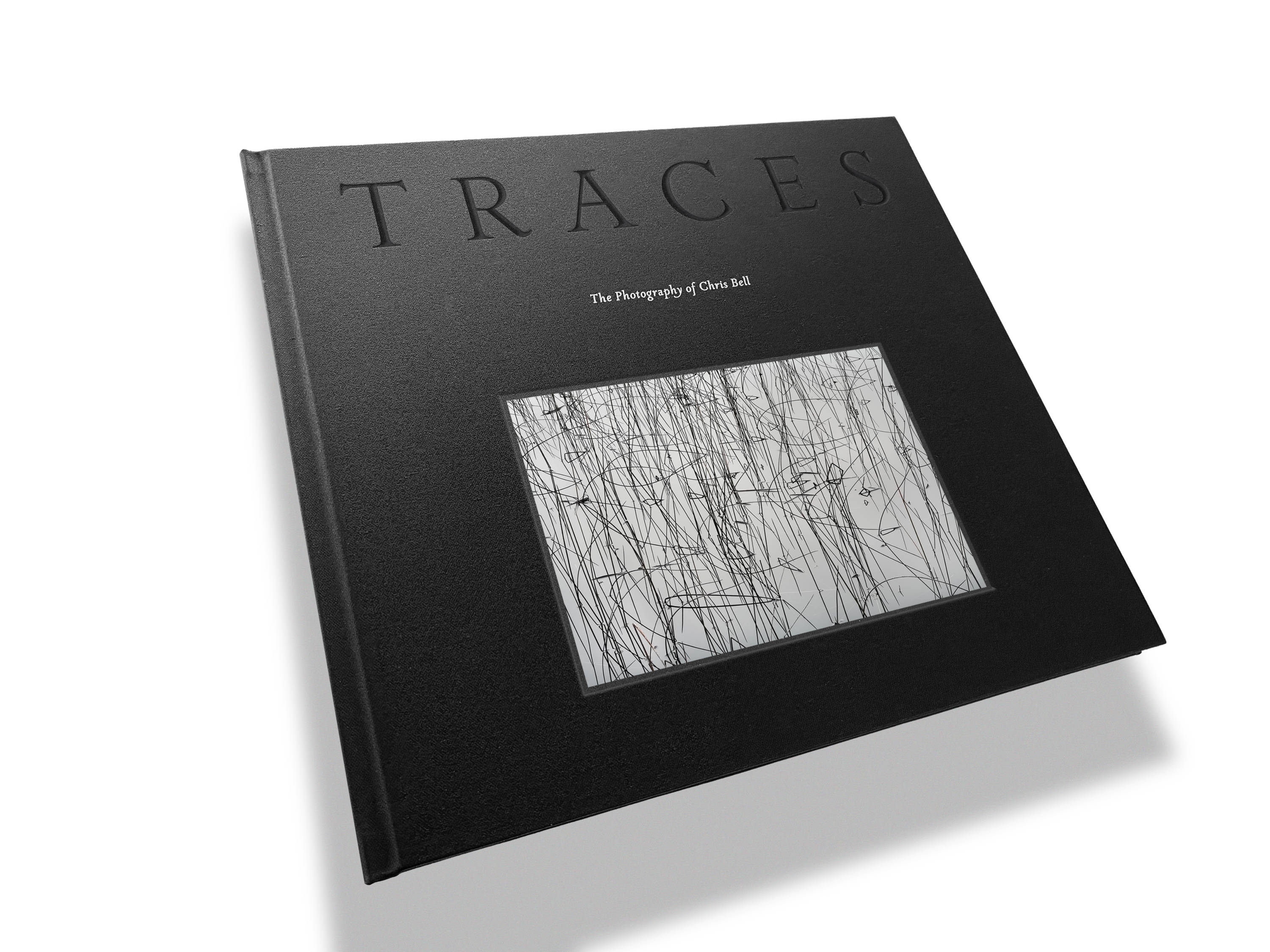 An image of the front cover of a large format book with a black cloth bound hard case with the title 'TRACES' in debossed black foil and a subtitle text that reads 'The Photography of Chris Bell' in silver foil. In the centre of the book cover is a debossed inlay image of fine pin rushes emerging from the water of a tarn.