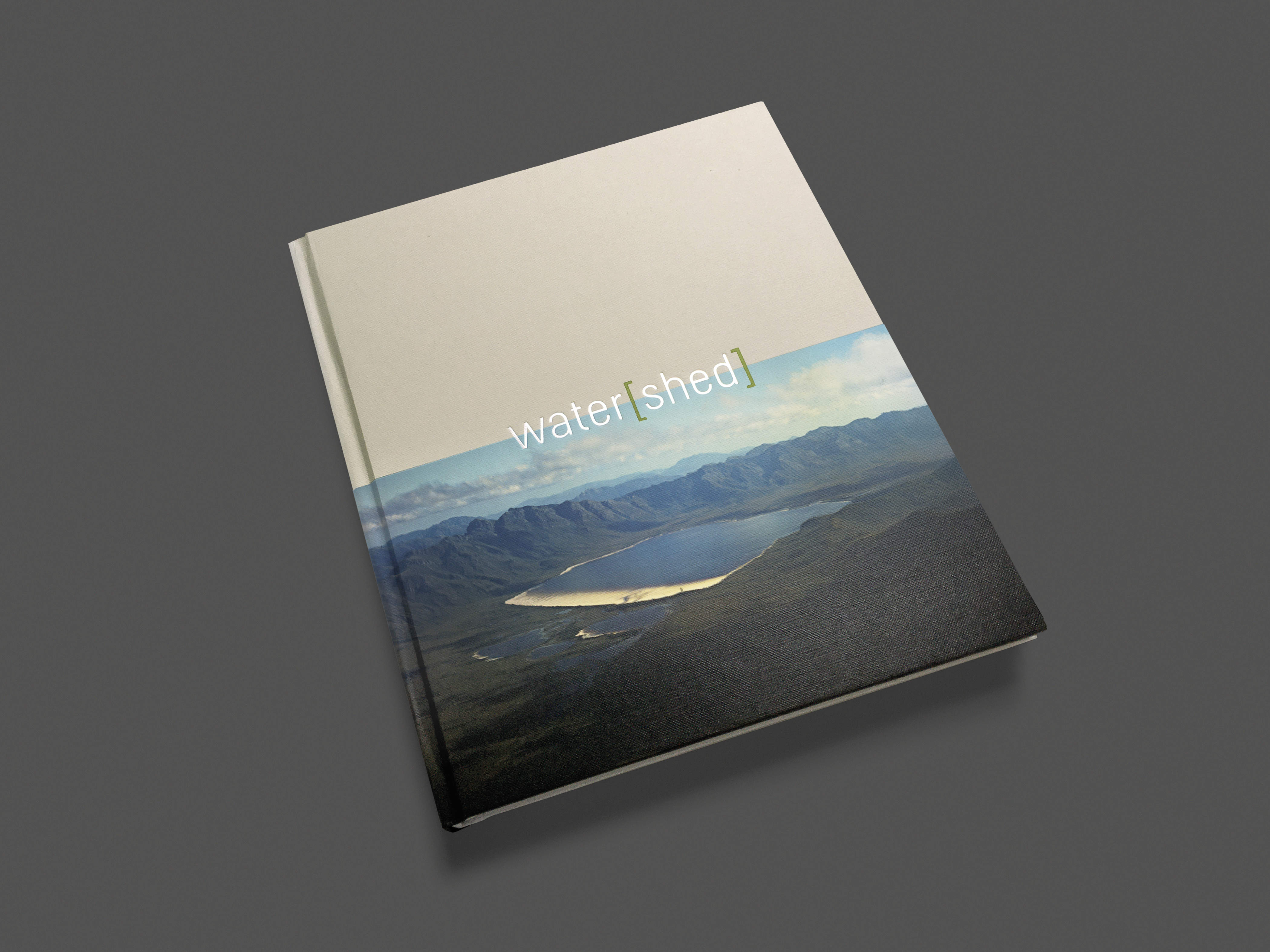 Image of the cover of the water[shed] exhibition publication. The title water[shed] sits in the middle of the cover and is debossed in silver foil and the word ‘shed’ is in bright green square brackets. The top half of the cover is a light grey colour. The bottom half of the cover features a photograph of Lake Pedder, Tasmania, c1970 taken by Wilf Elvey prior to the lake’s inundation by the Huon-Serpentine hydro-electric impoundment.
